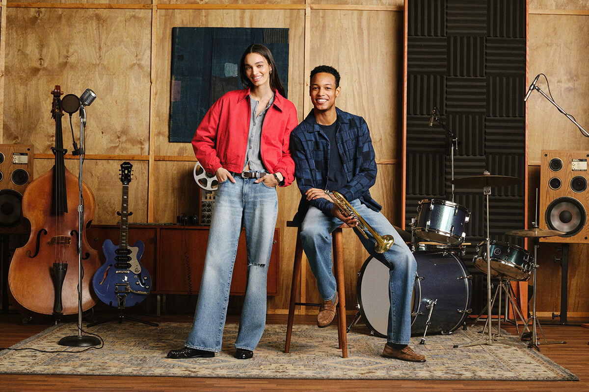 Two musicians posing in a music studio with instruments, including a cello, electric guitar, saxophone, and drum set. The woman wears a red jacket and light blue jeans, and the man wears a dark blue plaid shirt and jeans.