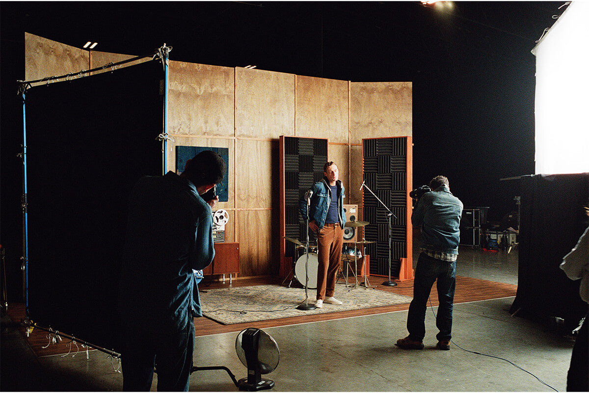 A behind-the-scenes look at a music recording studio setup, featuring a male artist standing in front of microphones and acoustic panels on a wooden set, with photographers and lighting equipment visible around him.