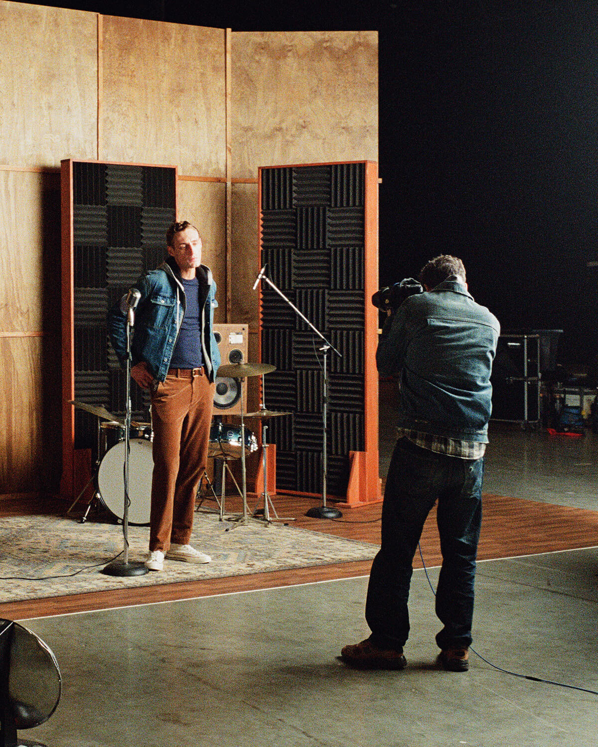 A man in a denim jacket and brown pants stands in a recording studio facing microphones, while another person in the foreground holds a camera. The background features wood-paneled walls, acoustic foam panels, and a drum kit.
