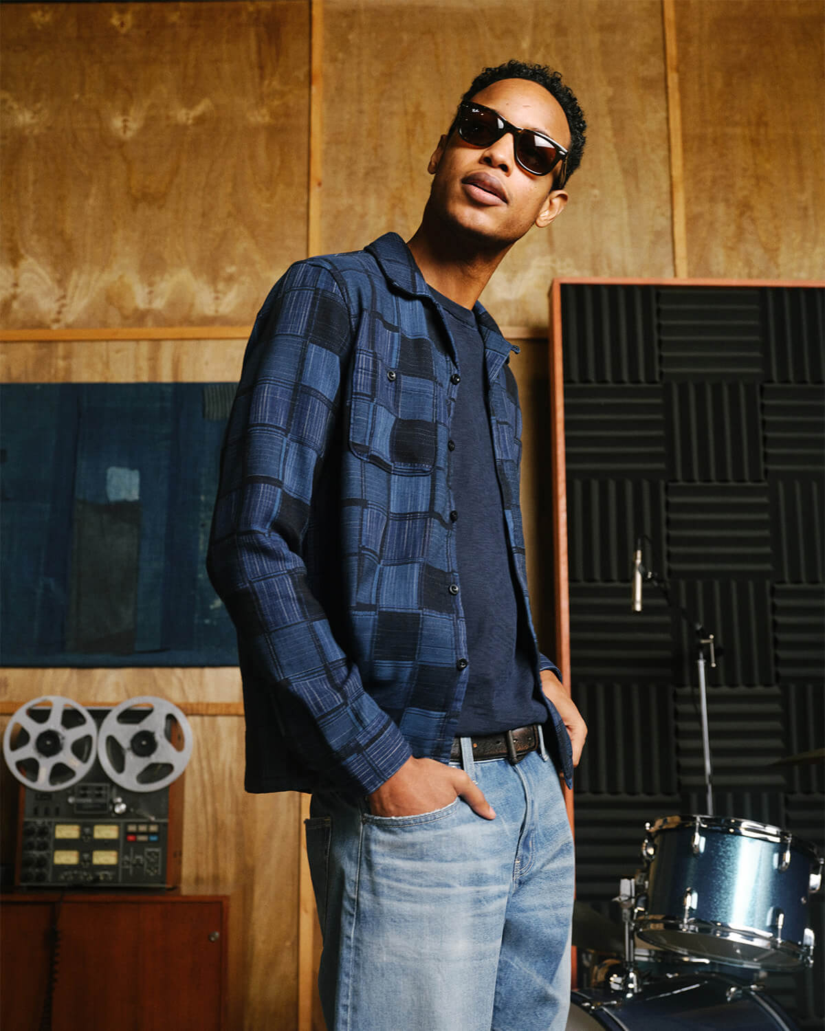 A man wearing a blue and black checkered flannel shirt over a dark t-shirt, paired with light blue jeans and sunglasses, standing in a music studio with wood-paneled walls, soundproofing foam, and musical equipment.