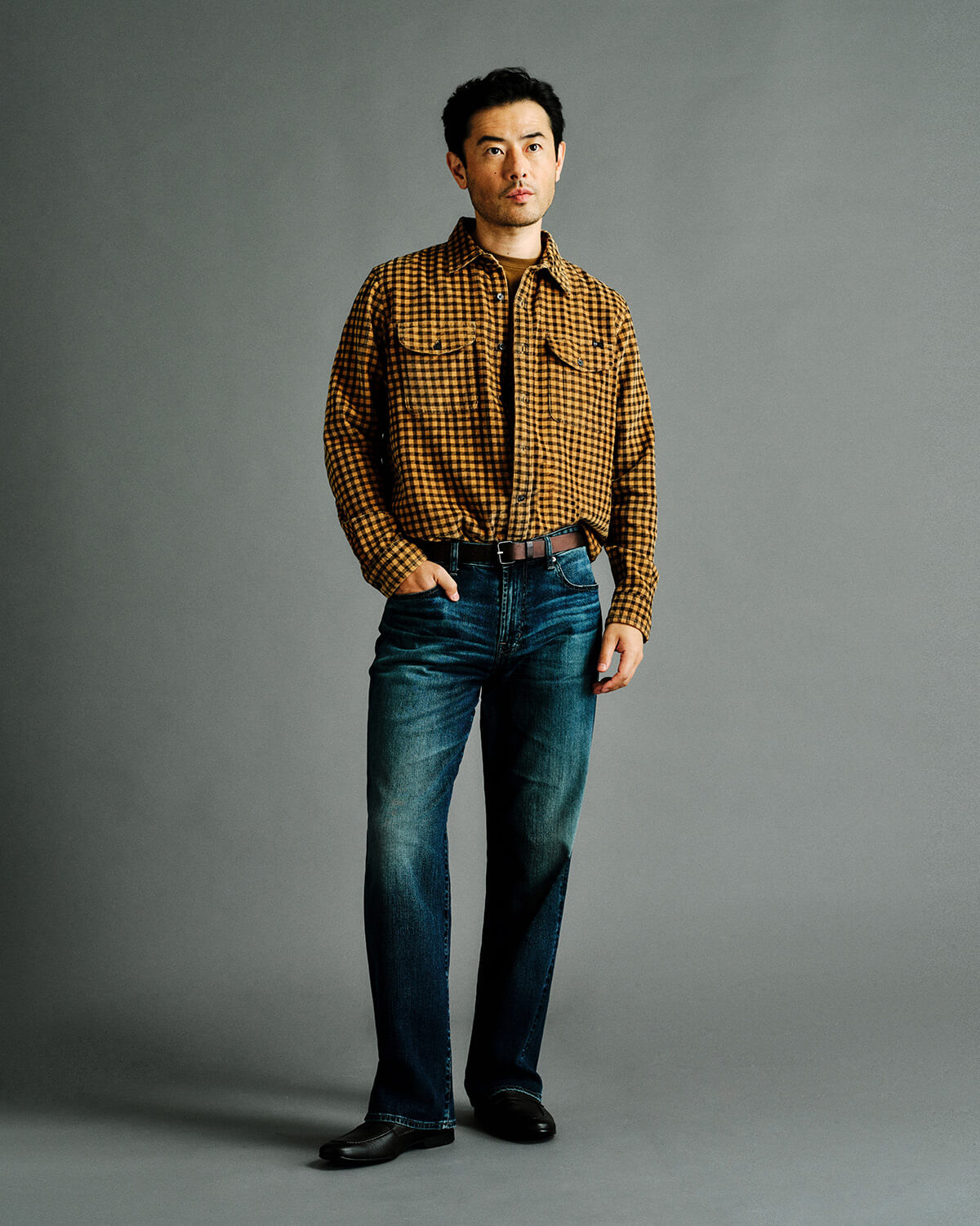 A man modeling a brown and black checkered long-sleeve button-down shirt with blue jeans, standing against a solid gray studio background.