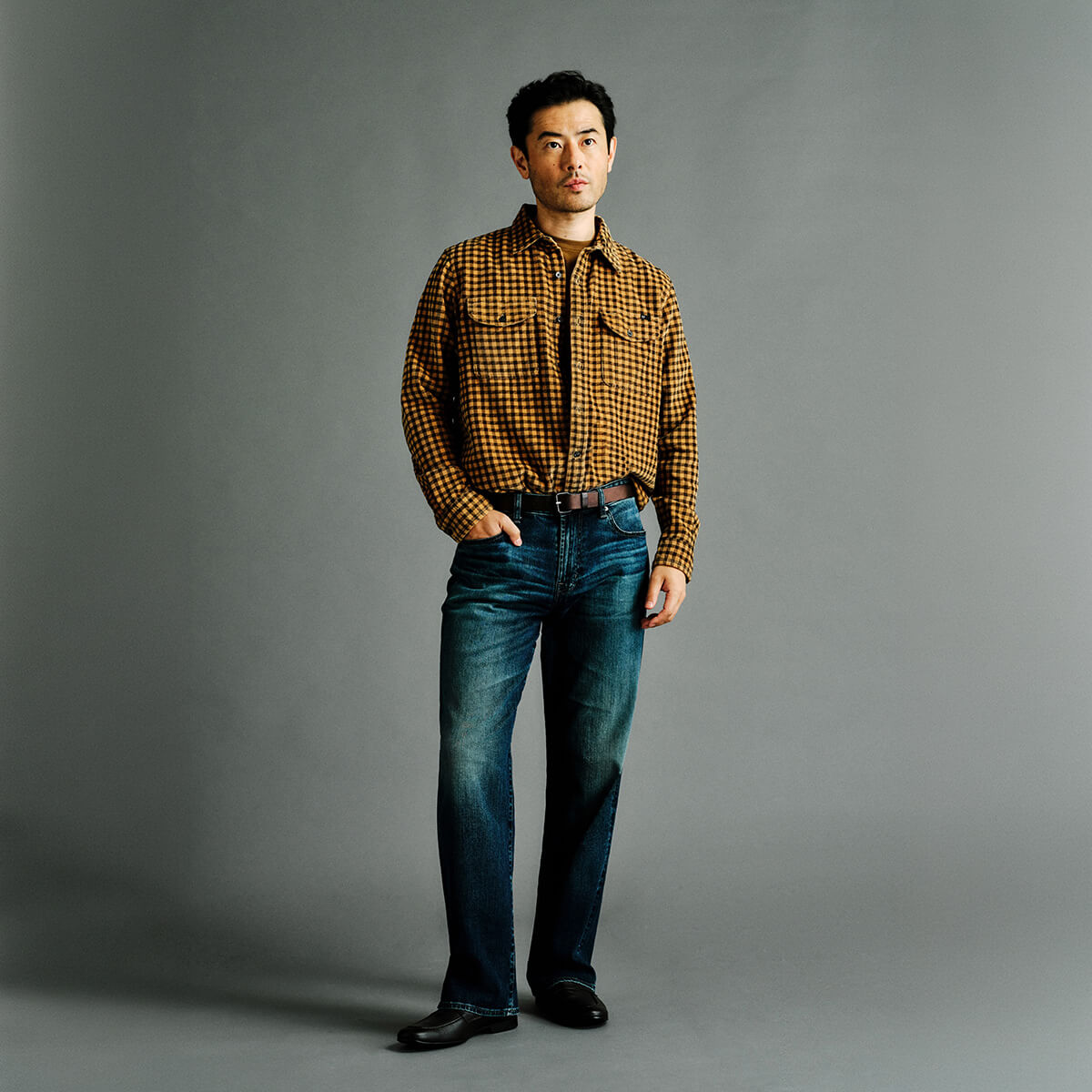 A man stands against a plain gray studio background wearing a brown and black checkered long-sleeve button-up shirt, dark blue jeans, and black shoes.