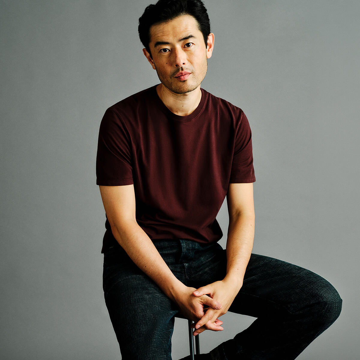 A man wearing a dark maroon crew-neck t-shirt and dark jeans, sitting against a solid gray studio background.