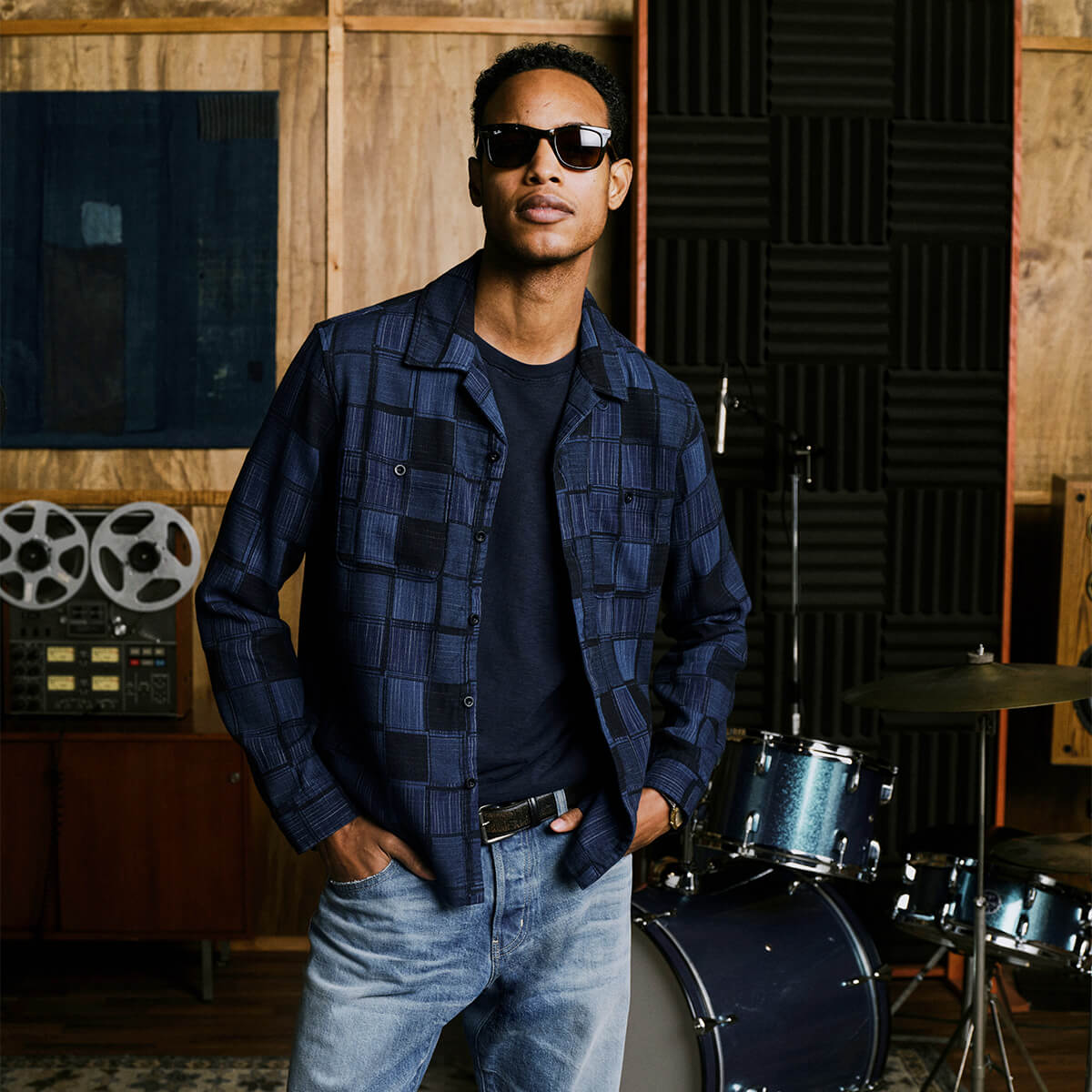 A man wearing a dark blue and black checkered button-up shirt over a black t-shirt, paired with light blue jeans and sunglasses, standing in a music studio with a drum kit and soundproofing panels in the background.