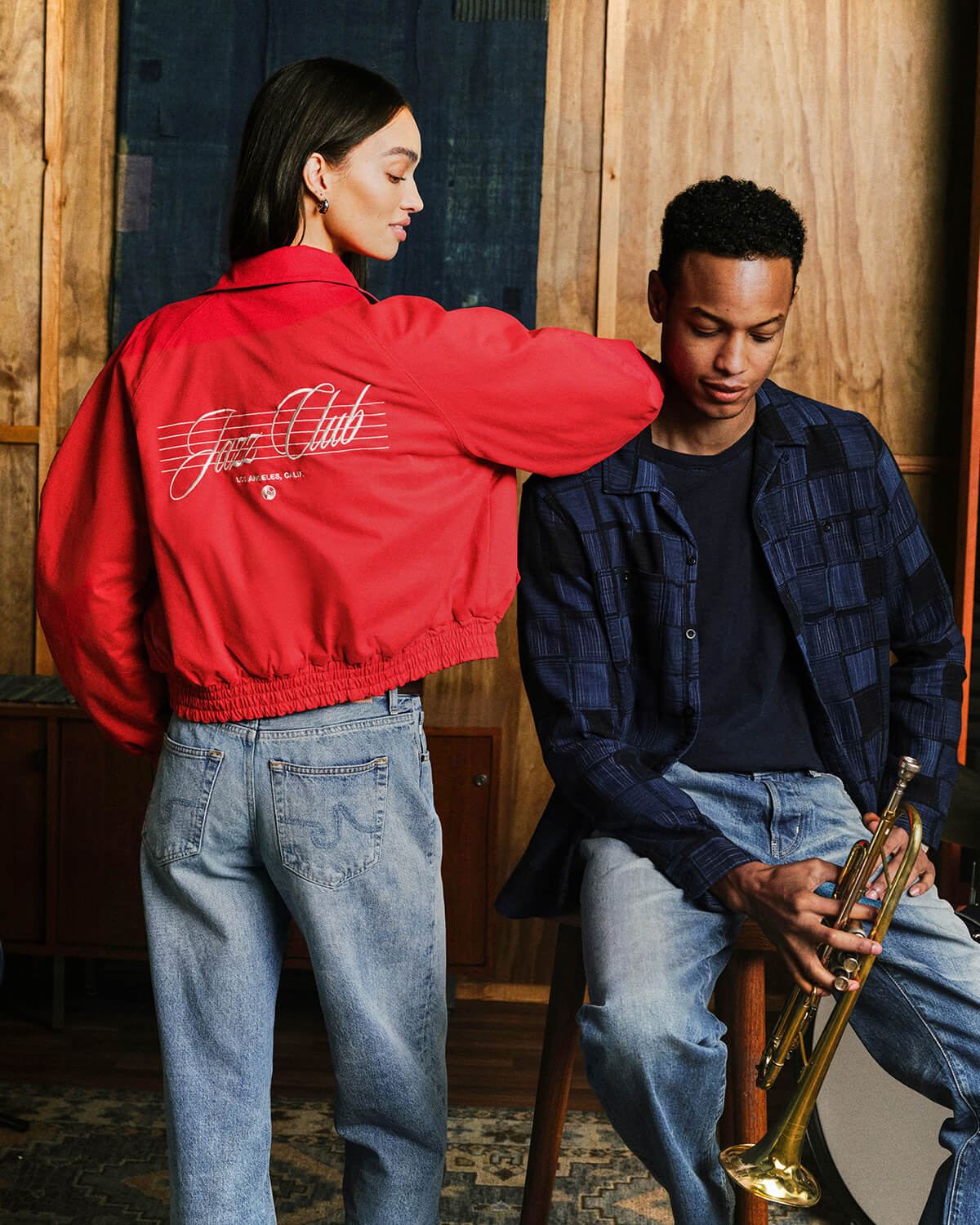 A model wearing a bright red bomber jacket with 'Just Club' printed in white script on the back, standing next to a man in a blue plaid shirt holding a trumpet in a rustic wood-paneled room.