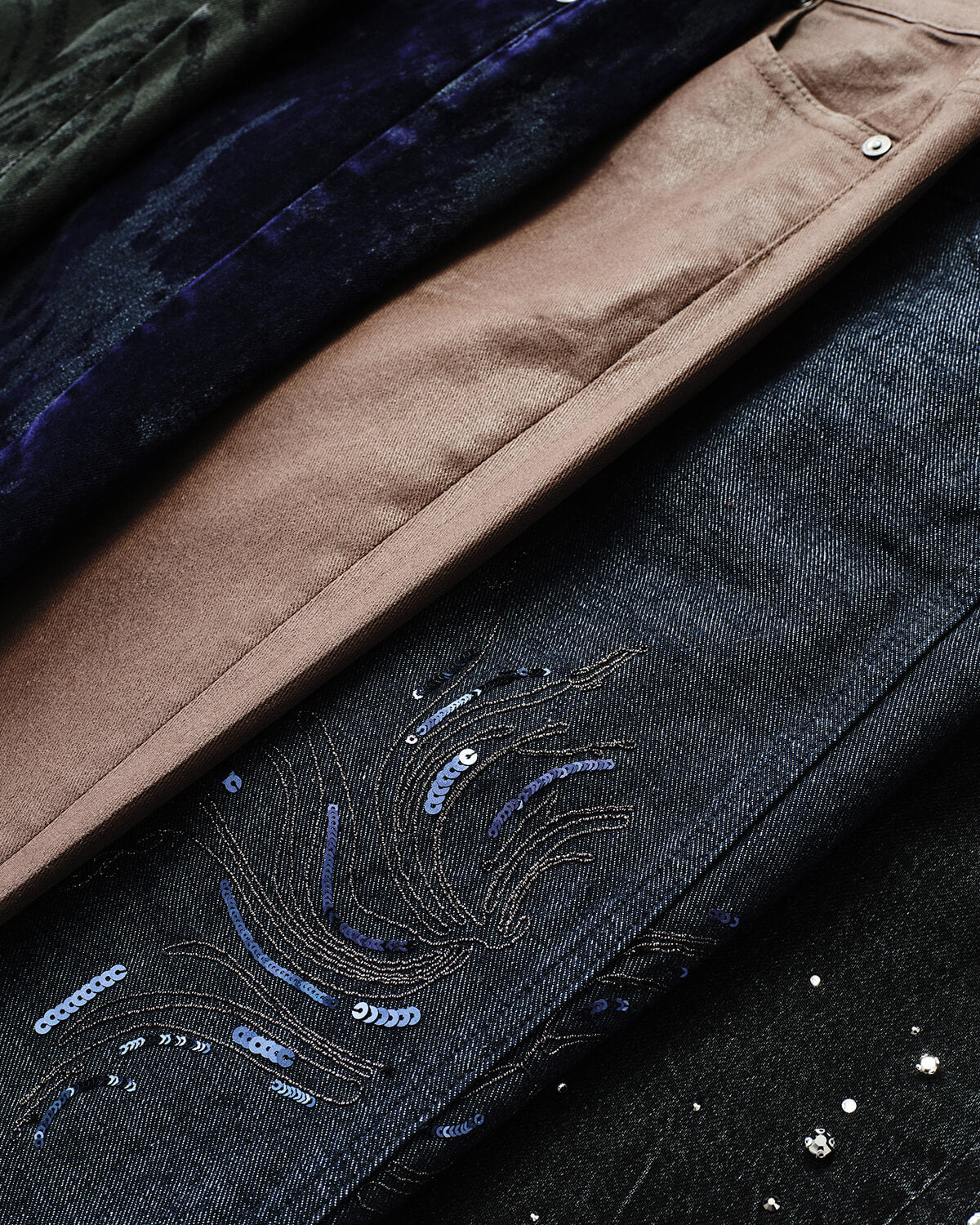 Close-up of folded denim garments in dark blue and black washes with embroidered wave-like sequin detailing and a tan leather trim.