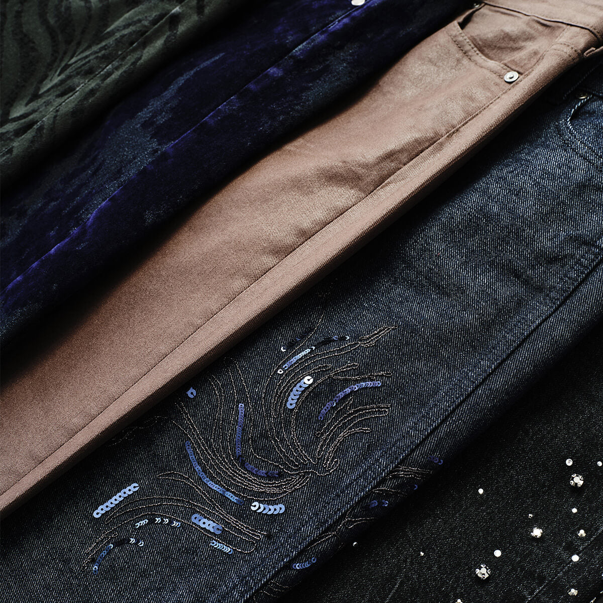 Close-up of folded denim jeans in multiple colors, including dark blue, brown, black, and a dark green velvet-like pair, with decorative sequin embroidery visible on the dark denim.