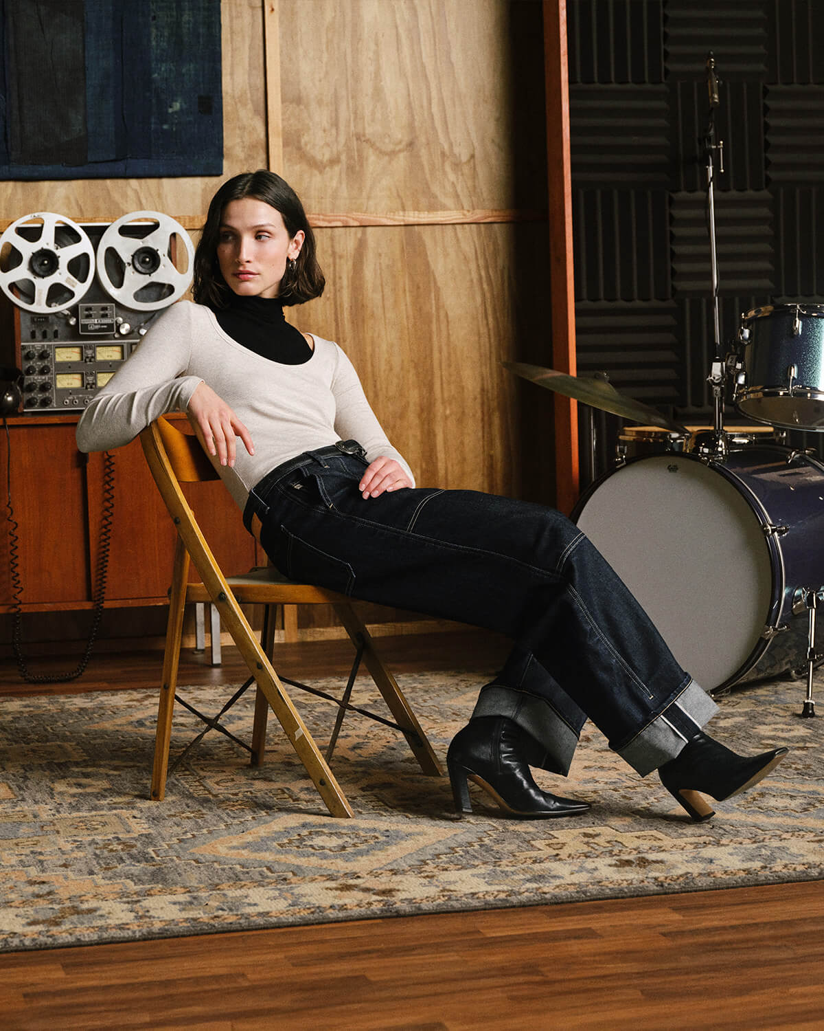 A woman sits on a wooden folding chair in a music studio, wearing a cream ribbed turtleneck, dark wide-leg jeans, and black ankle boots. A drum kit, acoustic wood wall, and vintage reel-to-reel machine are visible in the background.