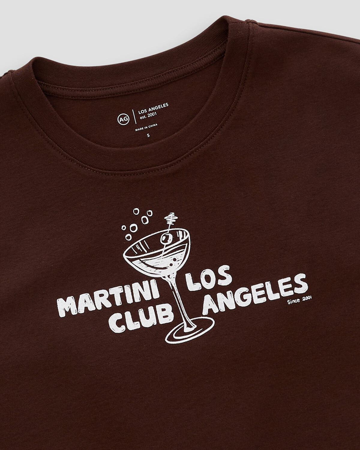 A close-up of a dark brown crewneck t-shirt featuring a white graphic print of a martini glass and the text 'MARTINI CLUB LOS ANGELES'.