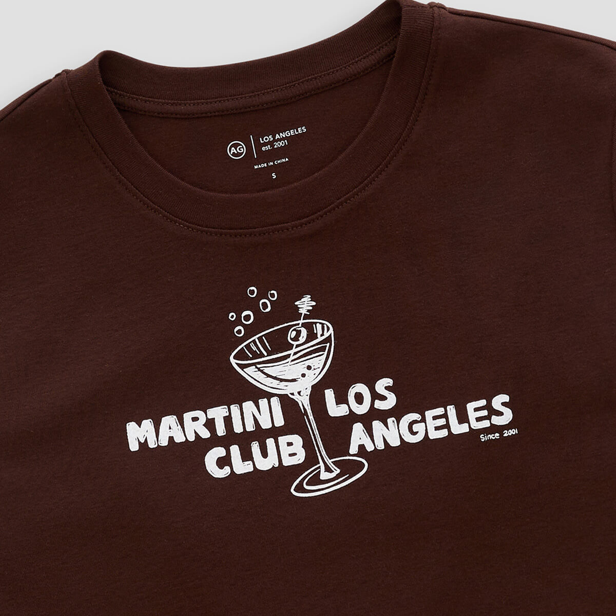 A close-up of a dark brown crewneck t-shirt featuring a white graphic print of a martini glass with the text 'MARTINI CLUB ANGELES'.