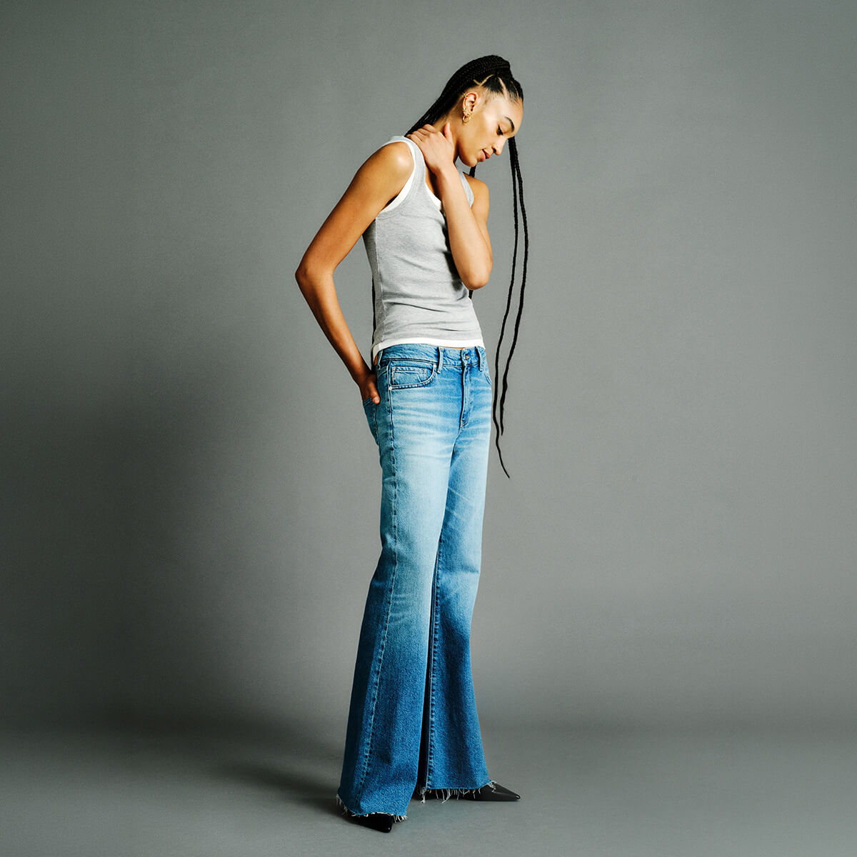 A woman modeling a pair of medium-wash blue flare jeans with a light gray ribbed tank top, standing against a solid gray studio background.
