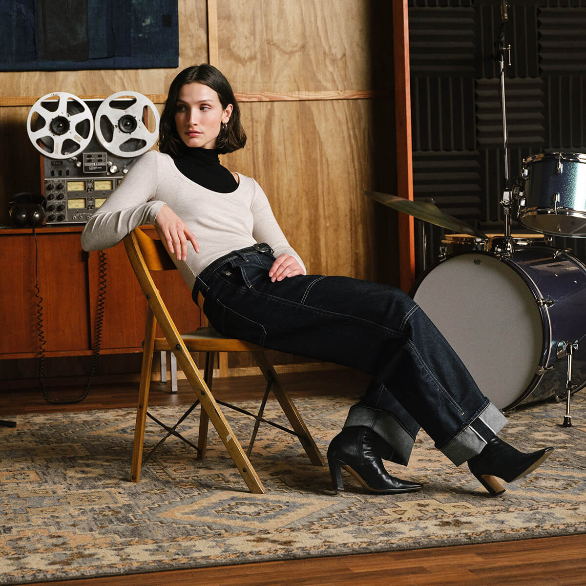 A woman sits on a wooden folding chair in a music studio setting, wearing a cream long-sleeve top, black turtleneck, dark blue jeans, and black ankle boots. A vintage reel-to-reel recorder, a drum set, and a patterned rug are visible in the background.