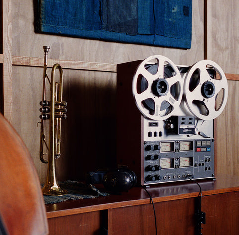 A vintage reel-to-reel tape recorder sits on a wooden console next to a brass trumpet, with a dark blue textile hanging on the wall behind it.