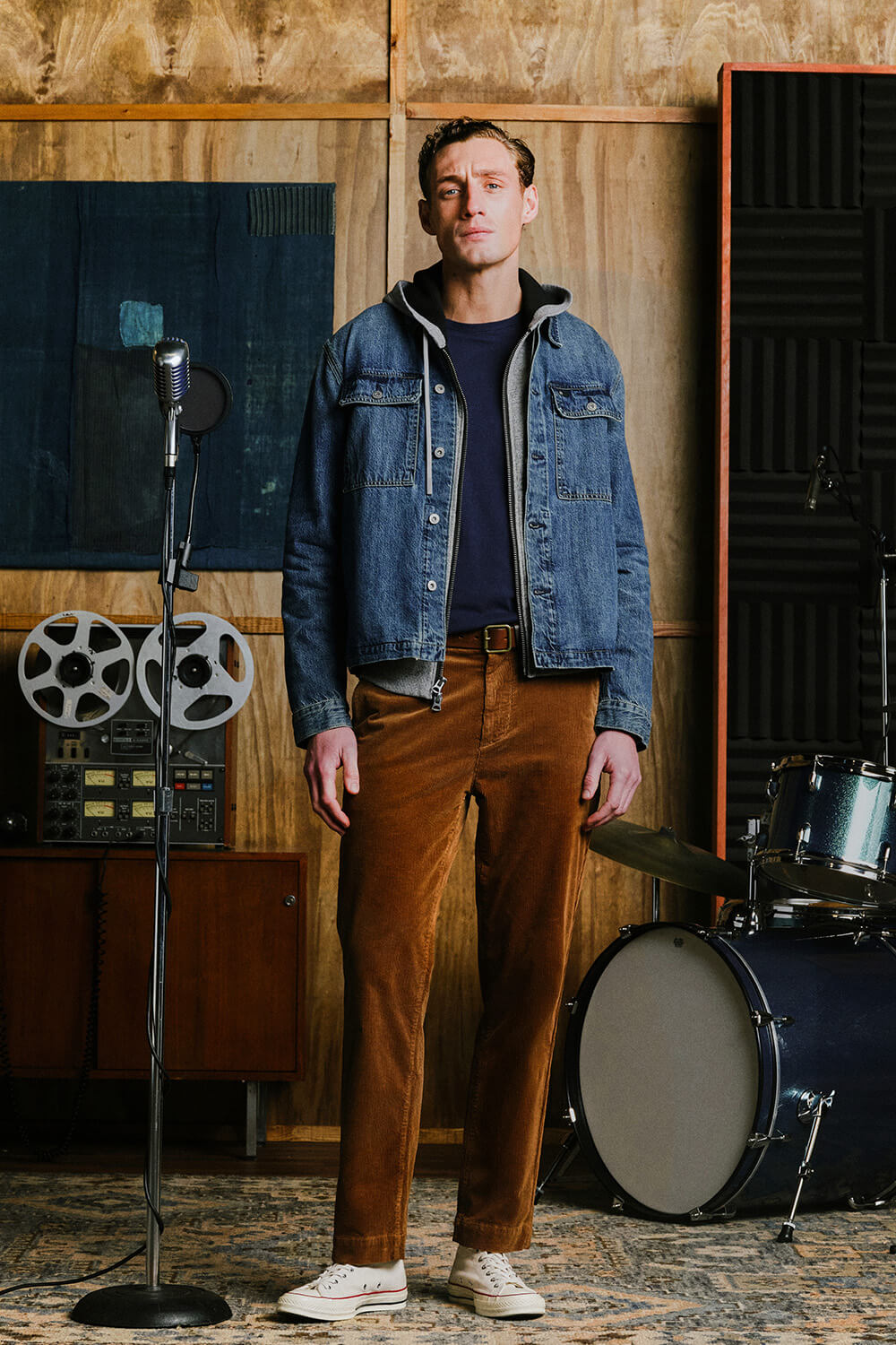A man stands in a music studio wearing a blue denim jacket over a dark navy shirt, brown corduroy trousers, and white sneakers. A vintage microphone stand, reel-to-reel machine, and drum set are visible in the background.