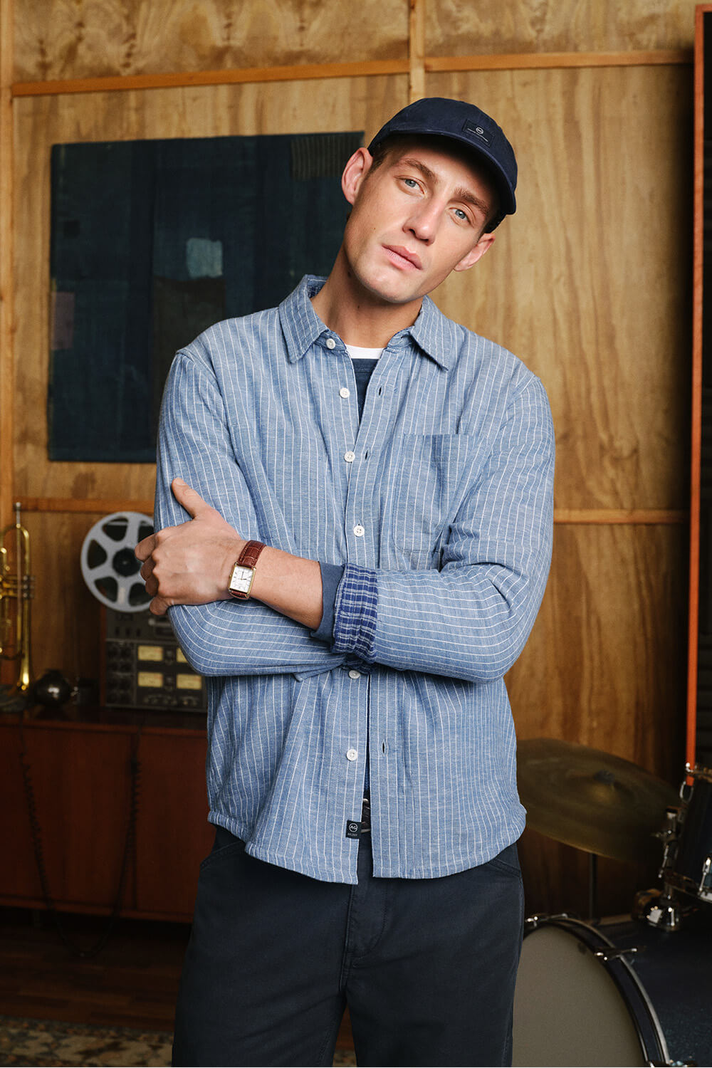 A man wearing a blue and white striped button-down shirt, dark pants, and a navy baseball cap stands with arms crossed in a studio setting with wood-paneled walls and musical equipment in the background.