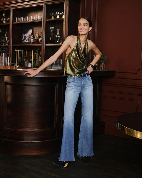 A woman posing in a dark, elegant room wearing a metallic olive green halter top and light-wash blue flared jeans.