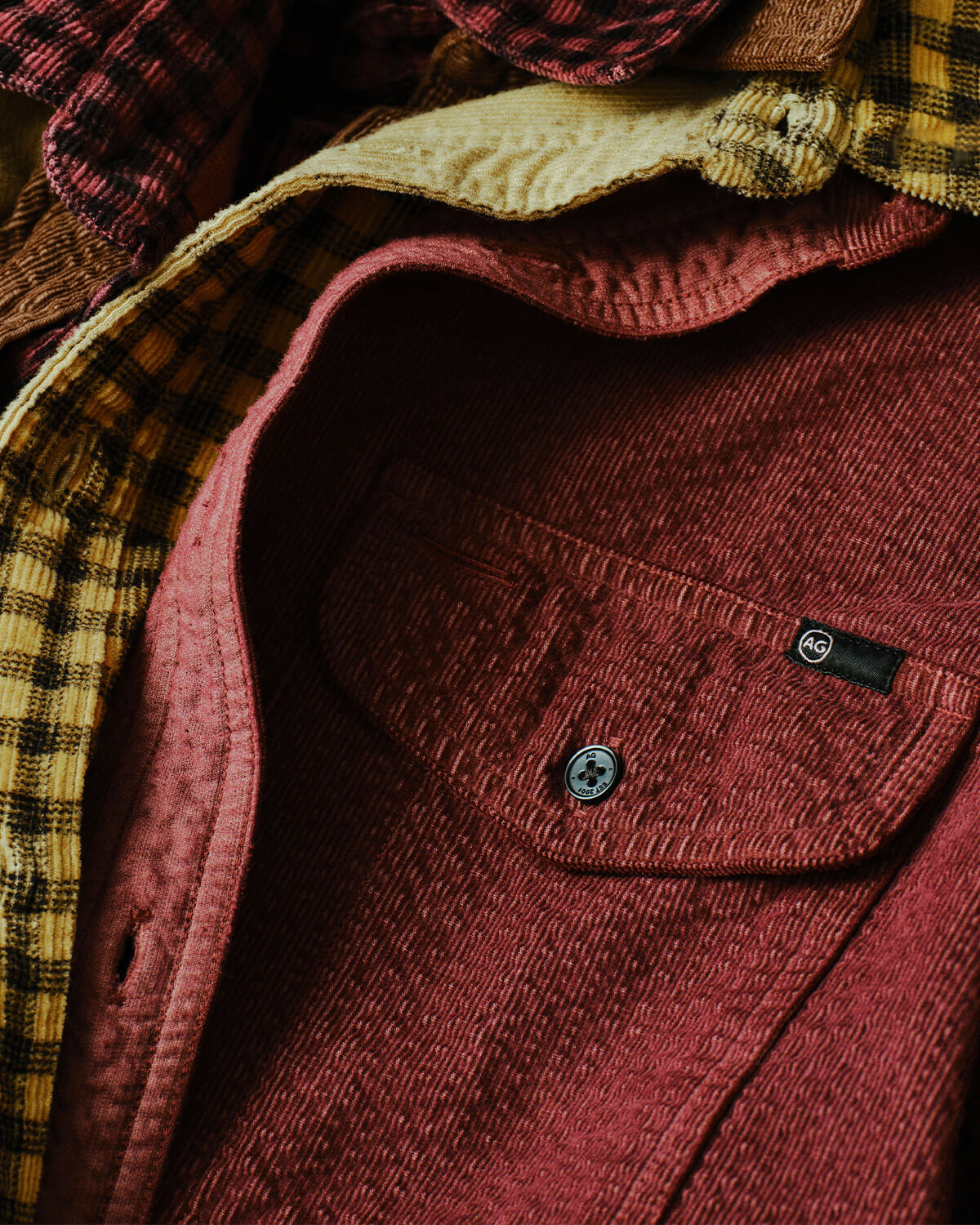 Close-up of a textured dusty rose corduroy overshirt with a chest pocket and snap button, layered over a yellow and black plaid flannel shirt.