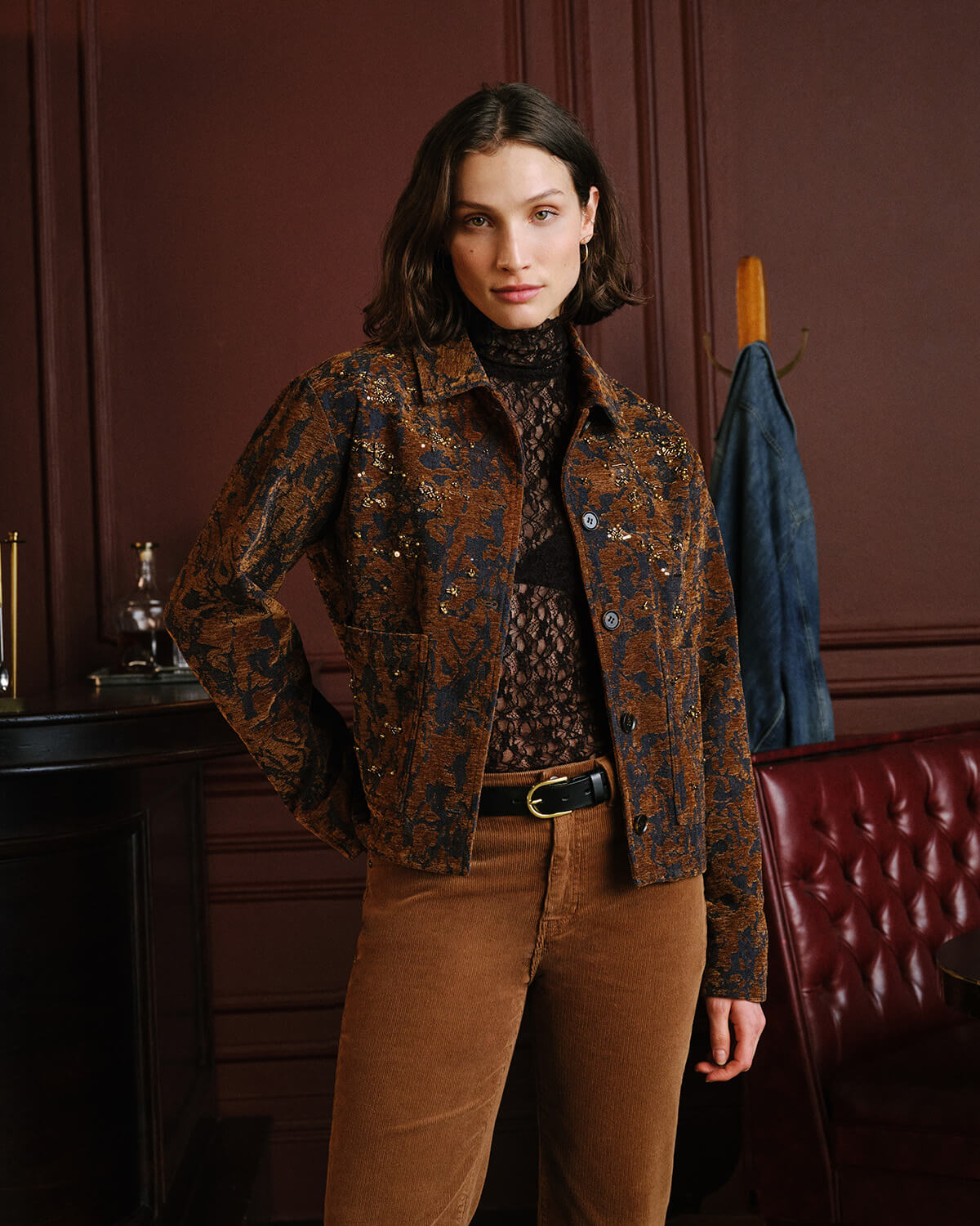 A woman models a brown and black patterned jacket with sequins, paired with brown corduroy trousers and a black lace top, standing in a dark, elegant room with a tufted red leather chair and dark wood paneling.