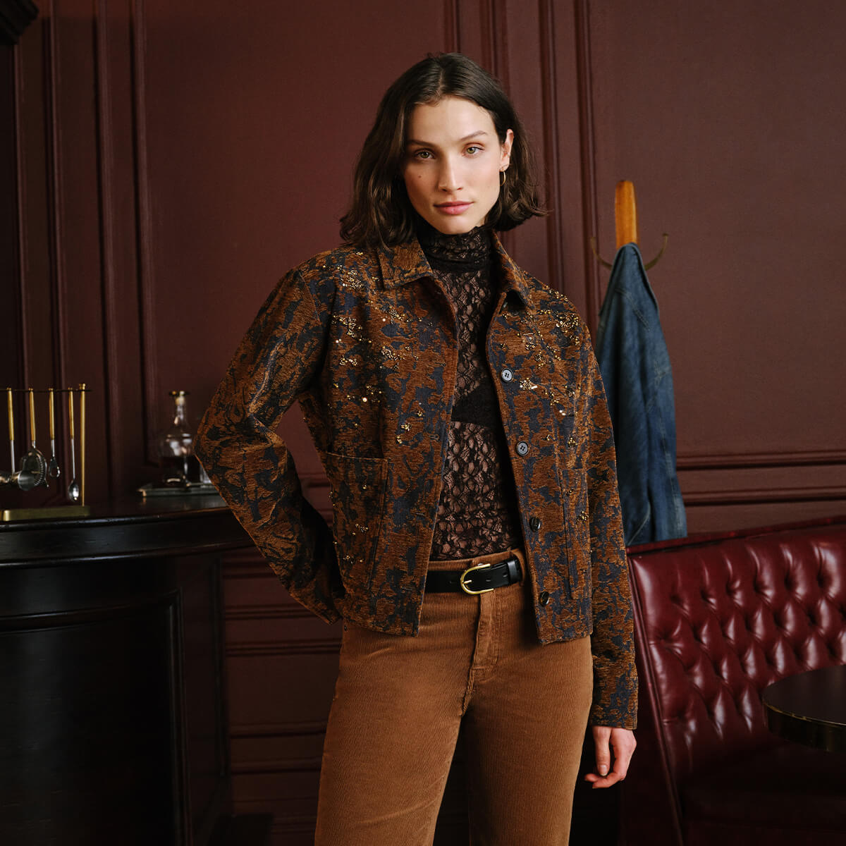 A woman stands in a dark, elegant room wearing a brown and black patterned jacket over a black lace turtleneck, paired with brown corduroy pants.