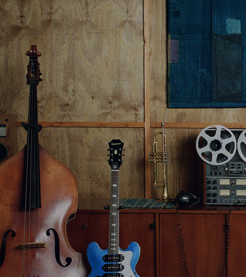 A blue electric guitar stands upright on a wooden cabinet in a room with plywood walls, alongside a double bass, a trumpet, and a reel-to-reel recording device.