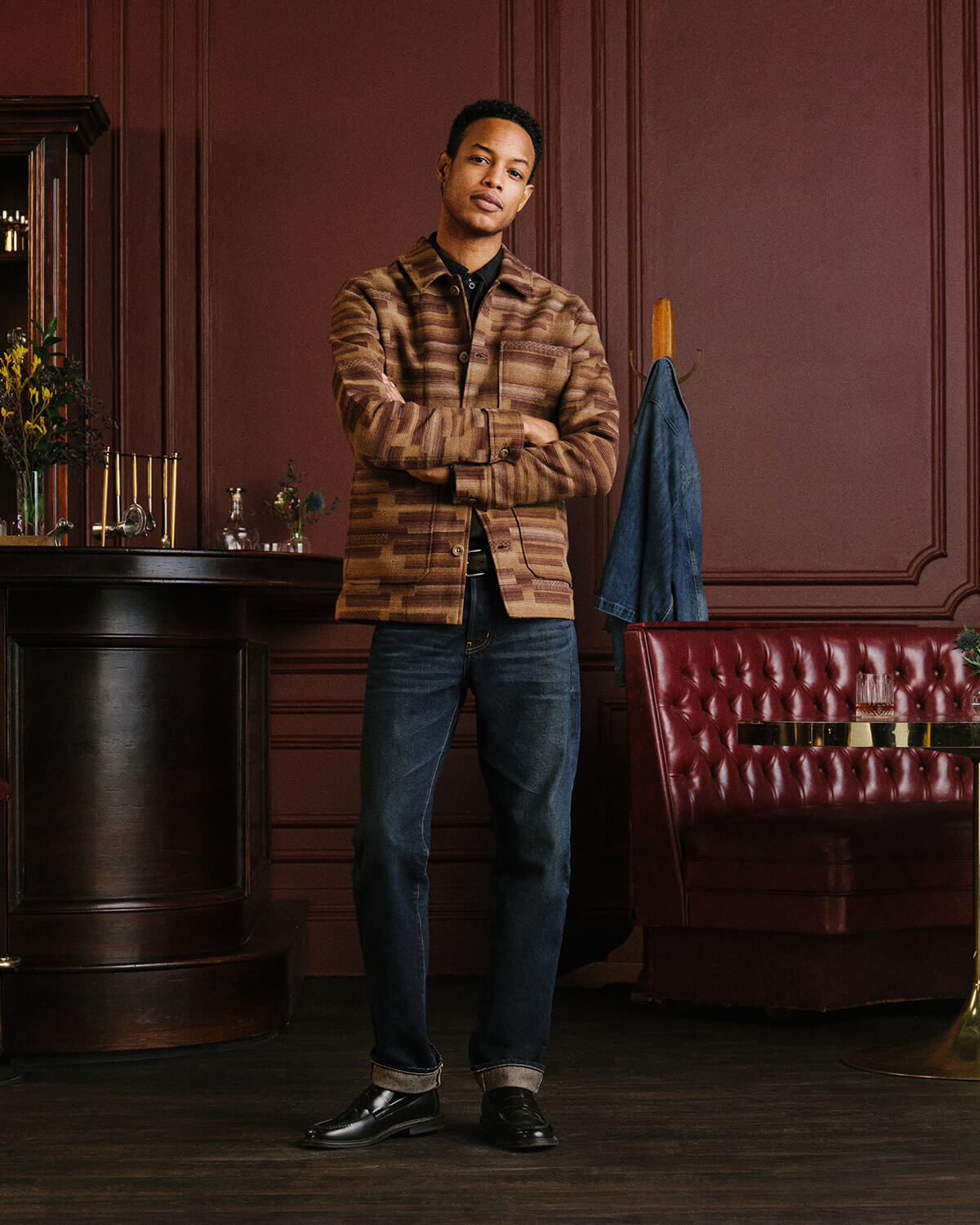 A man stands in a dimly lit, elegant room with dark wood paneling, wearing a brown and tan striped button-up shirt, dark blue jeans, and black dress shoes. A red tufted leather booth and a blue jacket hanging on a stand are visible in the background.