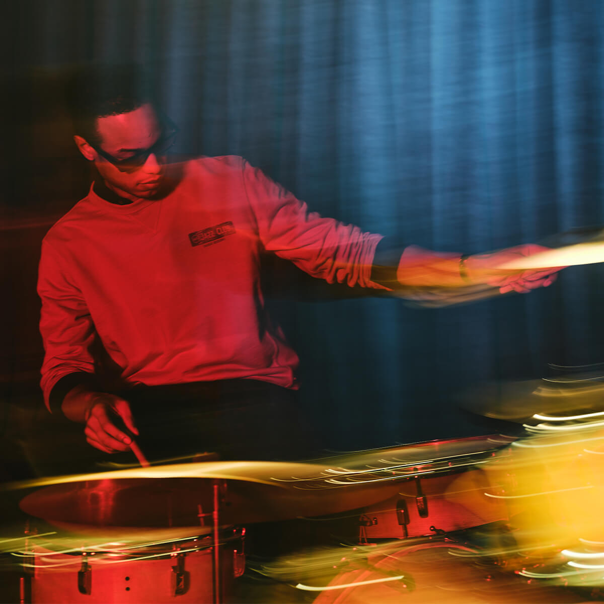 A drummer in a red long-sleeve shirt playing cymbals and drums, captured with motion blur and dramatic red and blue lighting against a dark curtain background.