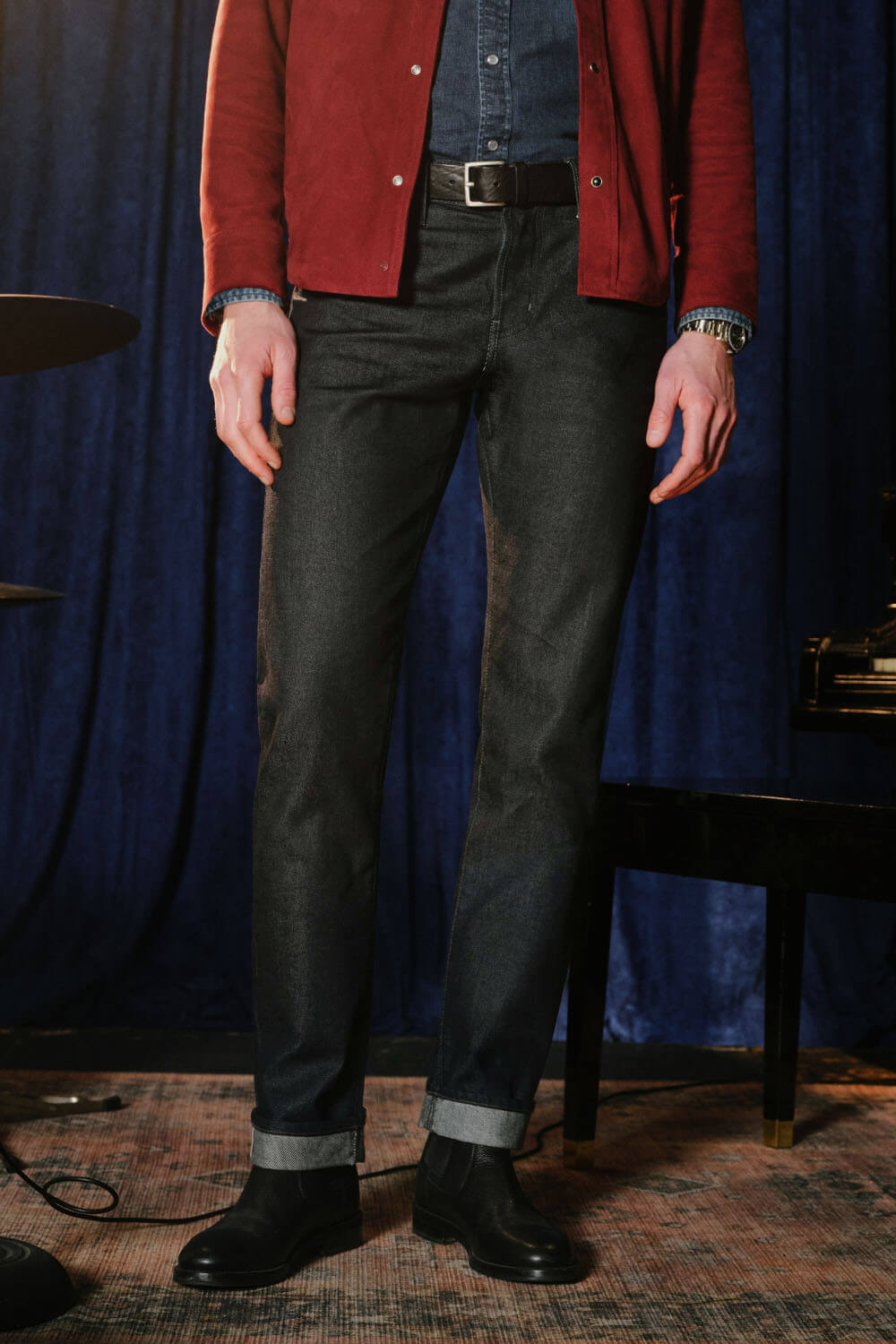 A person wearing black slim-fit jeans with a faded wash, styled with a red jacket, blue denim shirt, and black boots, standing in front of a dark blue velvet curtain.