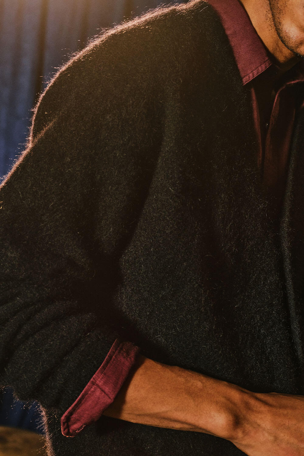 A close-up lifestyle shot of a person wearing a dark charcoal gray fuzzy knit cardigan over a burgundy collared shirt, with dramatic warm lighting highlighting the texture of the cardigan.