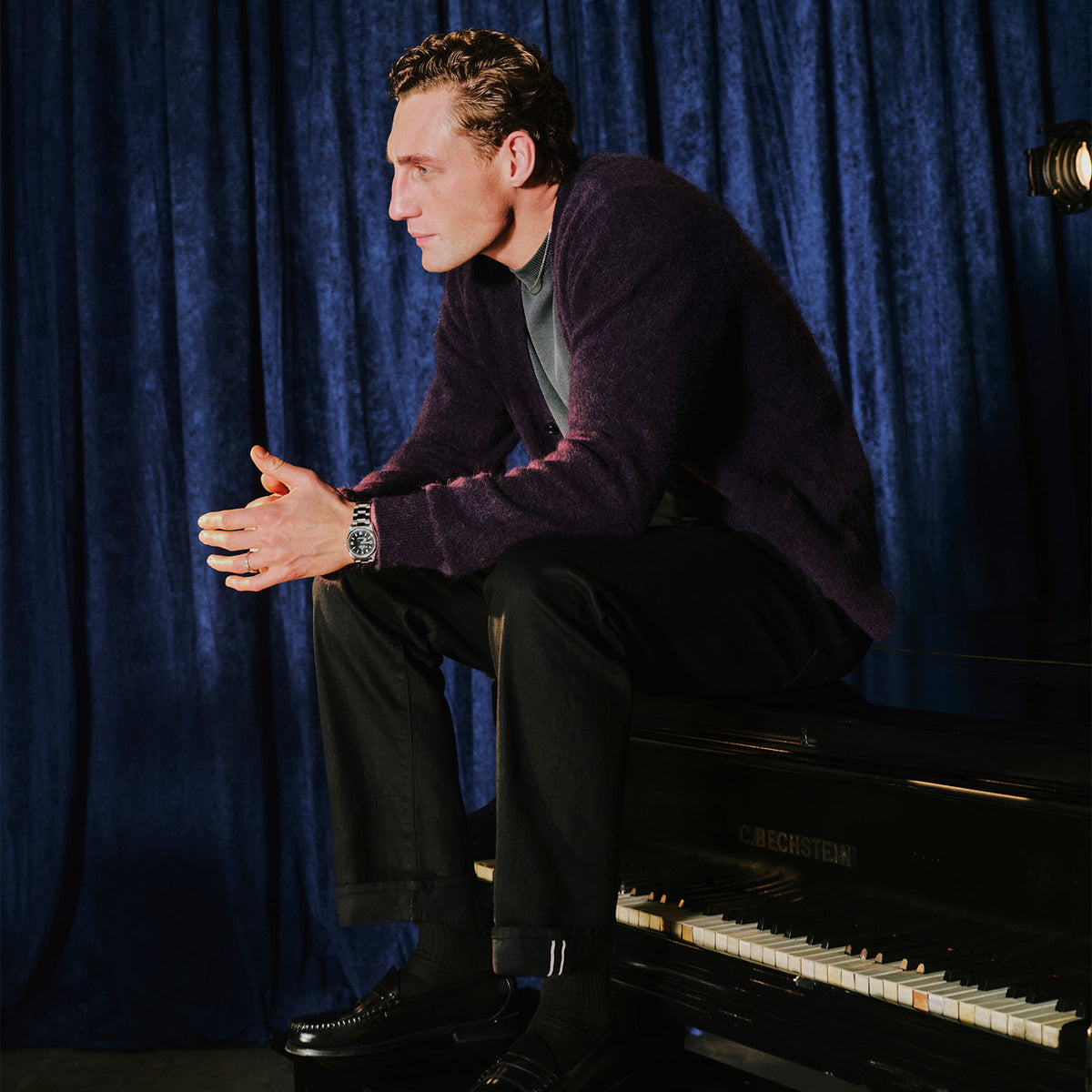 A man in a dark purple knit cardigan, black trousers, and black loafers sits on a black piano against a deep blue velvet curtain backdrop.
