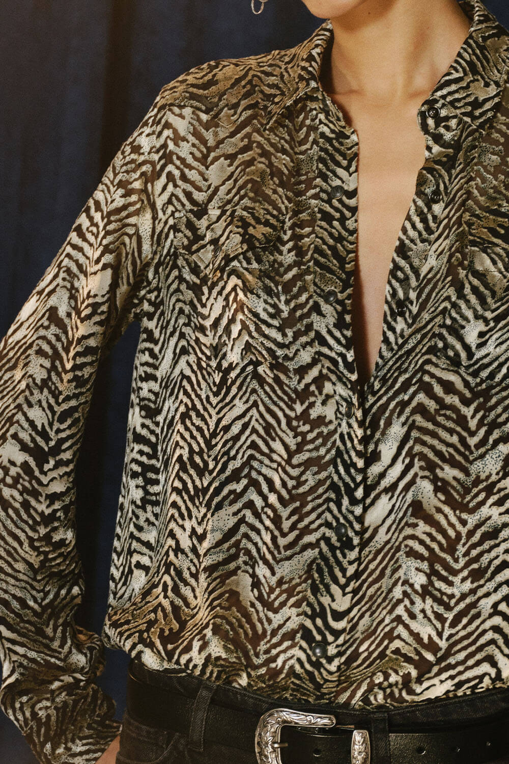 A close-up of a woman wearing a long-sleeve button-up blouse with a textured zebra-like animal print in brown, beige, and black. The blouse is paired with a black belt with a silver buckle.