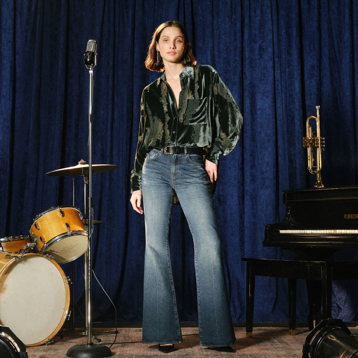 A model stands in a studio setting wearing a dark green velvet long-sleeve blouse with a floral print and blue flare jeans. The background features a deep blue velvet curtain, with musical props including a piano, trumpet, and a drum kit on the left.