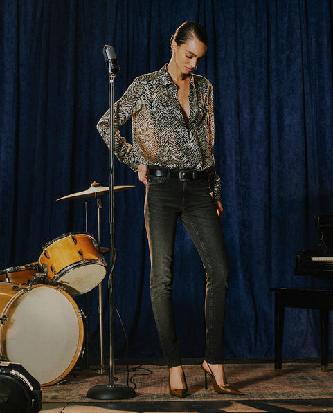A woman stands in a studio setting wearing a long-sleeve black and beige patterned blouse, black skinny jeans with side slit details, and brown pointed-toe heels. She is posed in front of a blue velvet curtain with a microphone stand, a drum kit, and a piano visible.