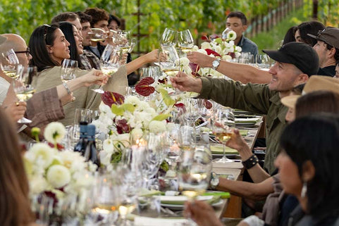 Group of people enjoying a meal together at an outdoor event with wine glasses and floral arrangements.