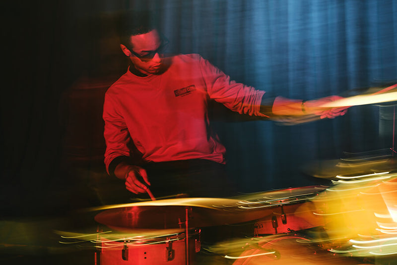 A drummer in a red long-sleeve shirt playing cymbals and drums on stage under dramatic red and blue lighting, with motion blur across the foreground.
