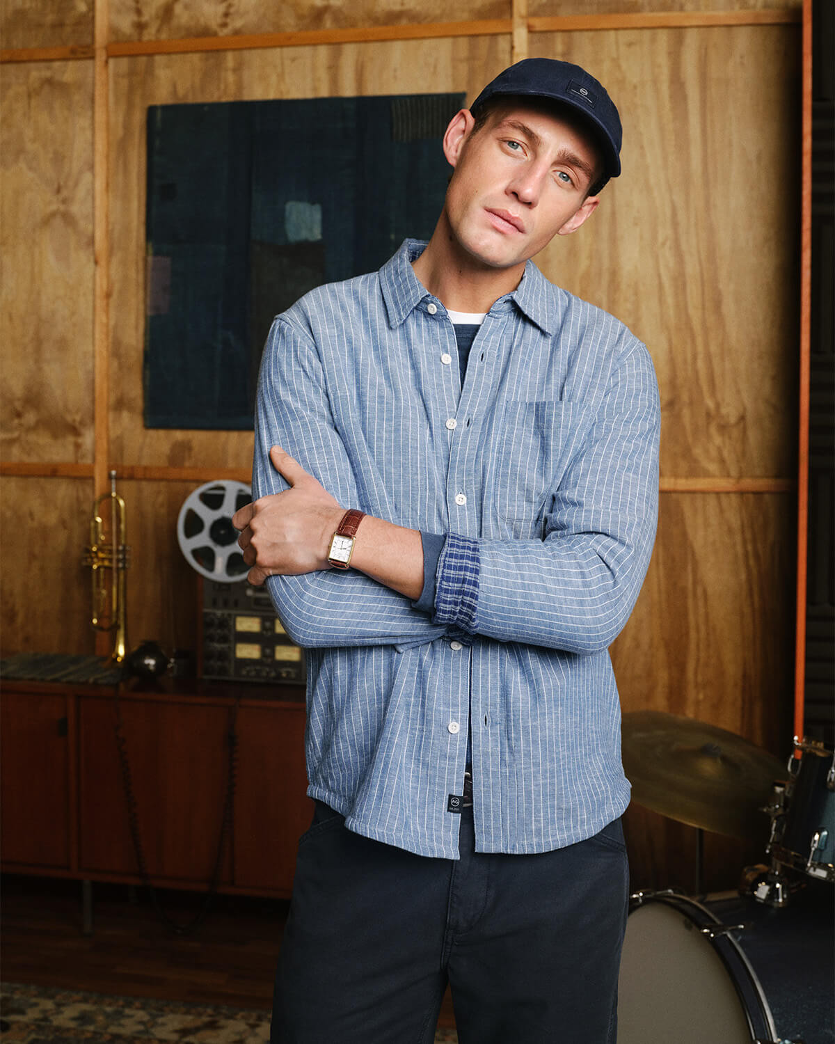 A man wearing a light blue and white vertically striped button-down shirt, dark pants, a dark baseball cap, and a watch, standing with arms crossed in front of a wood-paneled wall.