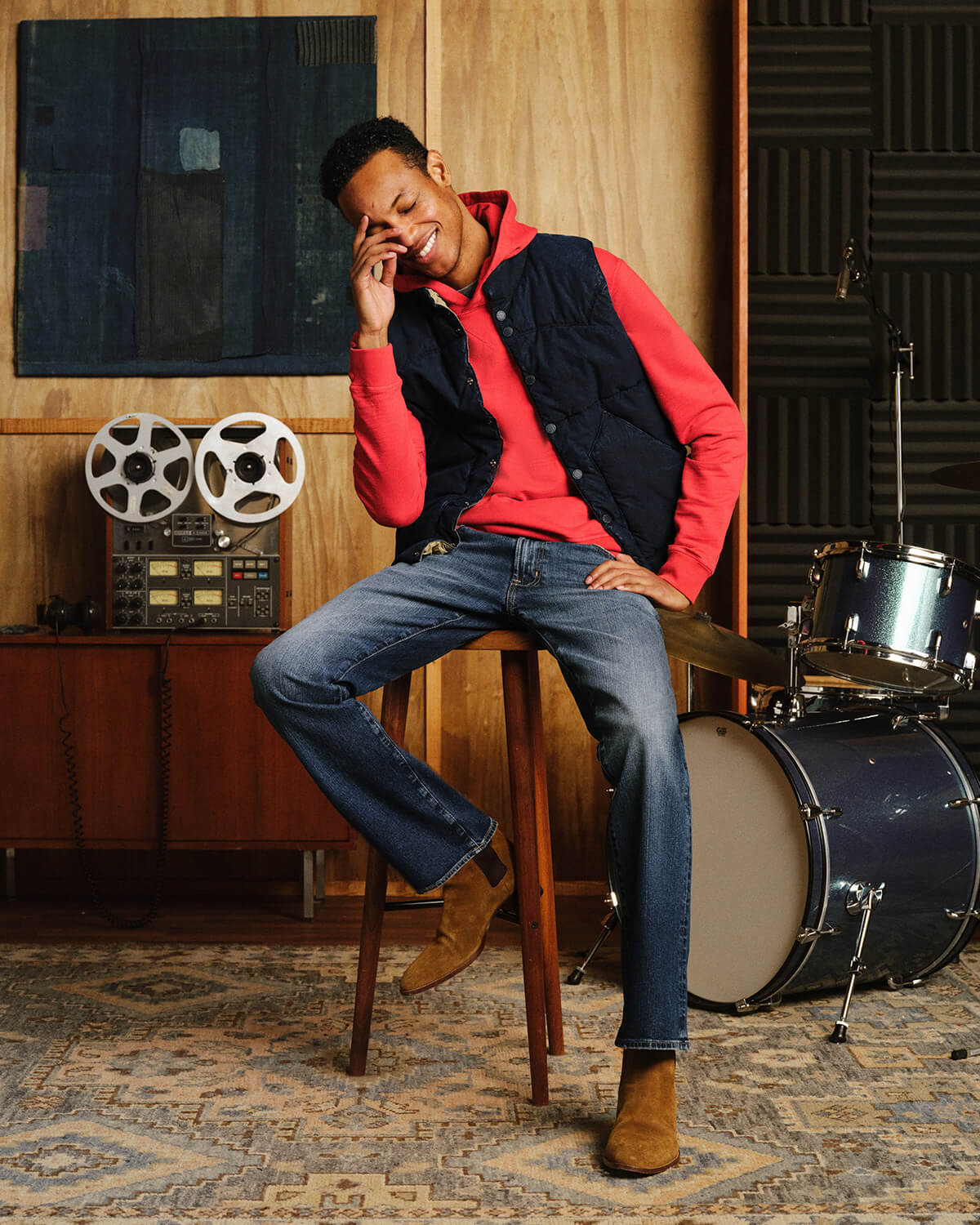 A man sitting on a wooden stool in a music studio setting, wearing a red long-sleeve shirt under a navy quilted vest, blue jeans, and brown boots. A reel-to-reel machine and a drum set are visible in the background.