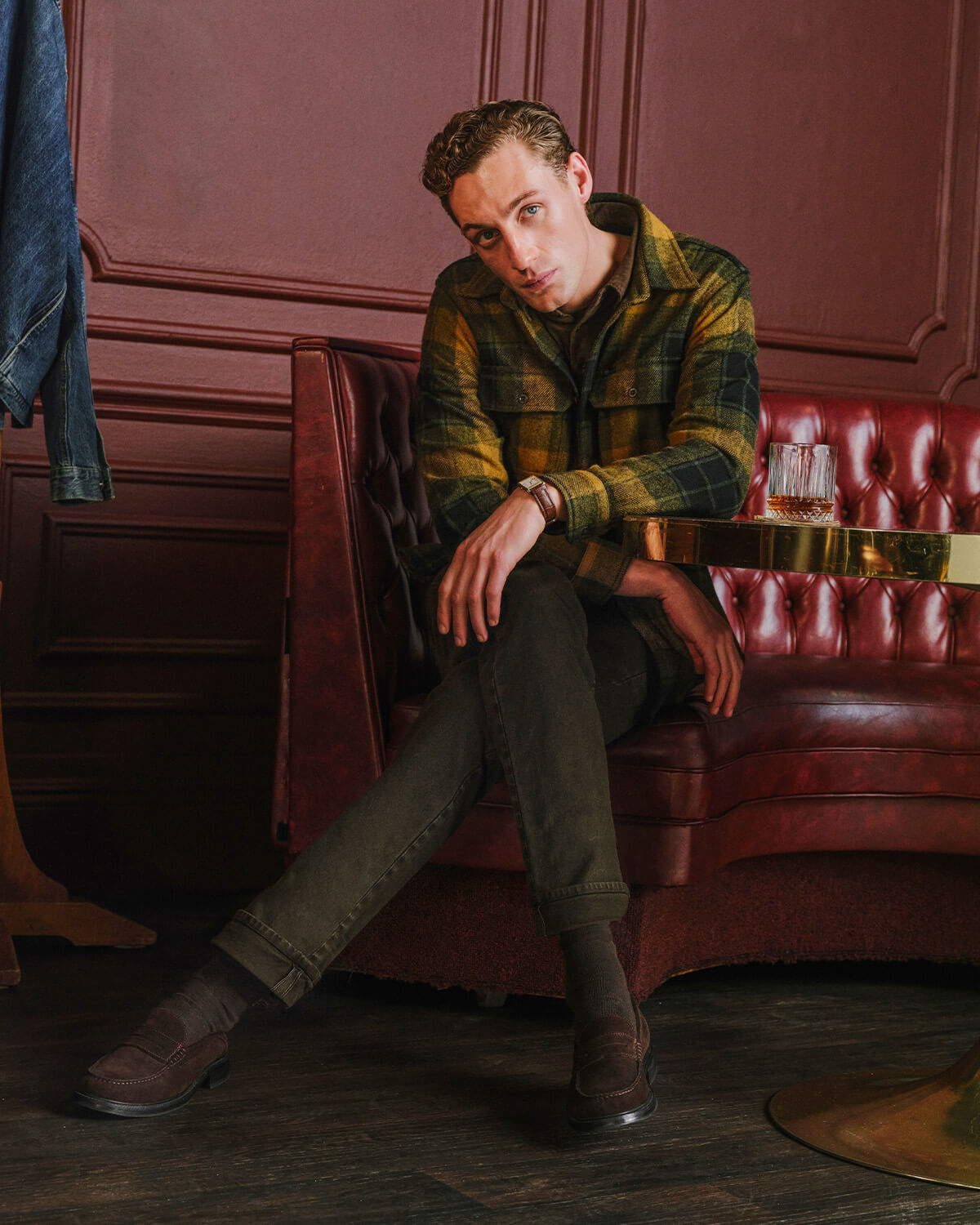 A man wearing a green and black plaid flannel shirt, dark olive trousers, and brown suede boots sits on a red leather tufted sofa in a dimly lit room with dark wood paneling.