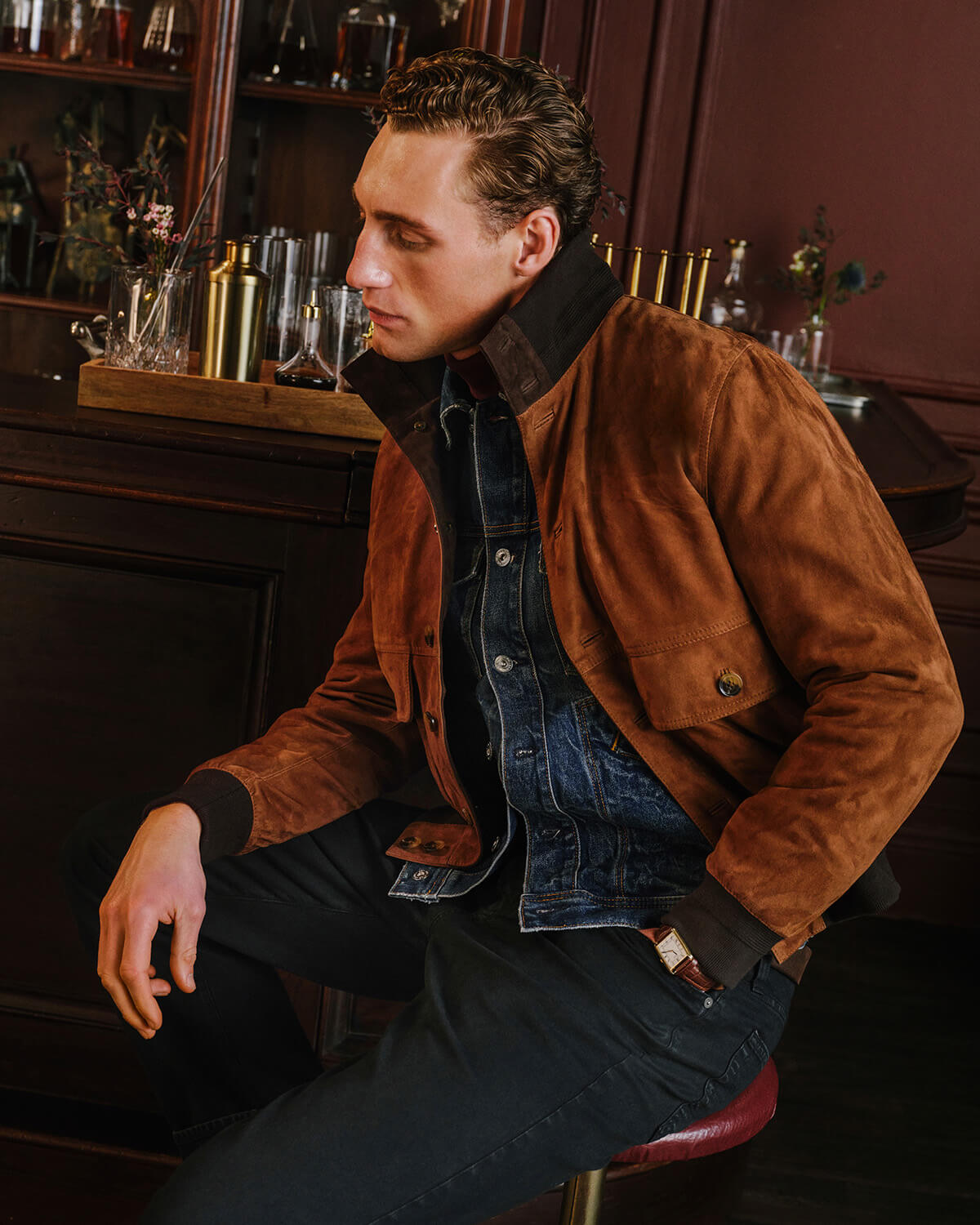 A man wearing a brown suede jacket over a blue denim shirt and black pants, seated on a stool in a dimly lit bar environment.
