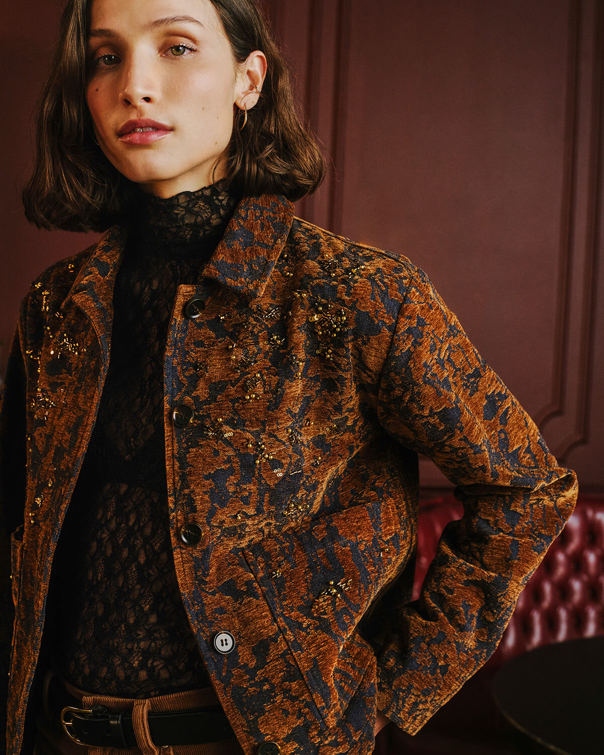 A woman wearing a brown and black patterned jacket over a black lace top, posed against a dark reddish-brown paneled wall.