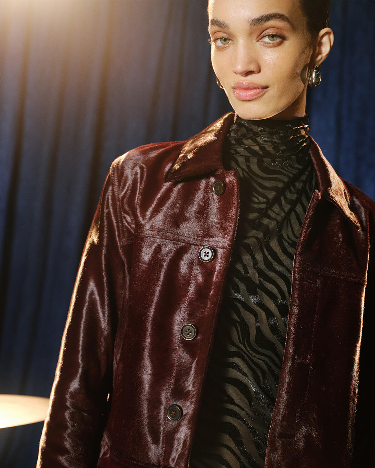 A model wearing a dark burgundy velvet jacket over a black and gray patterned turtleneck, photographed against a dark blue curtain background.