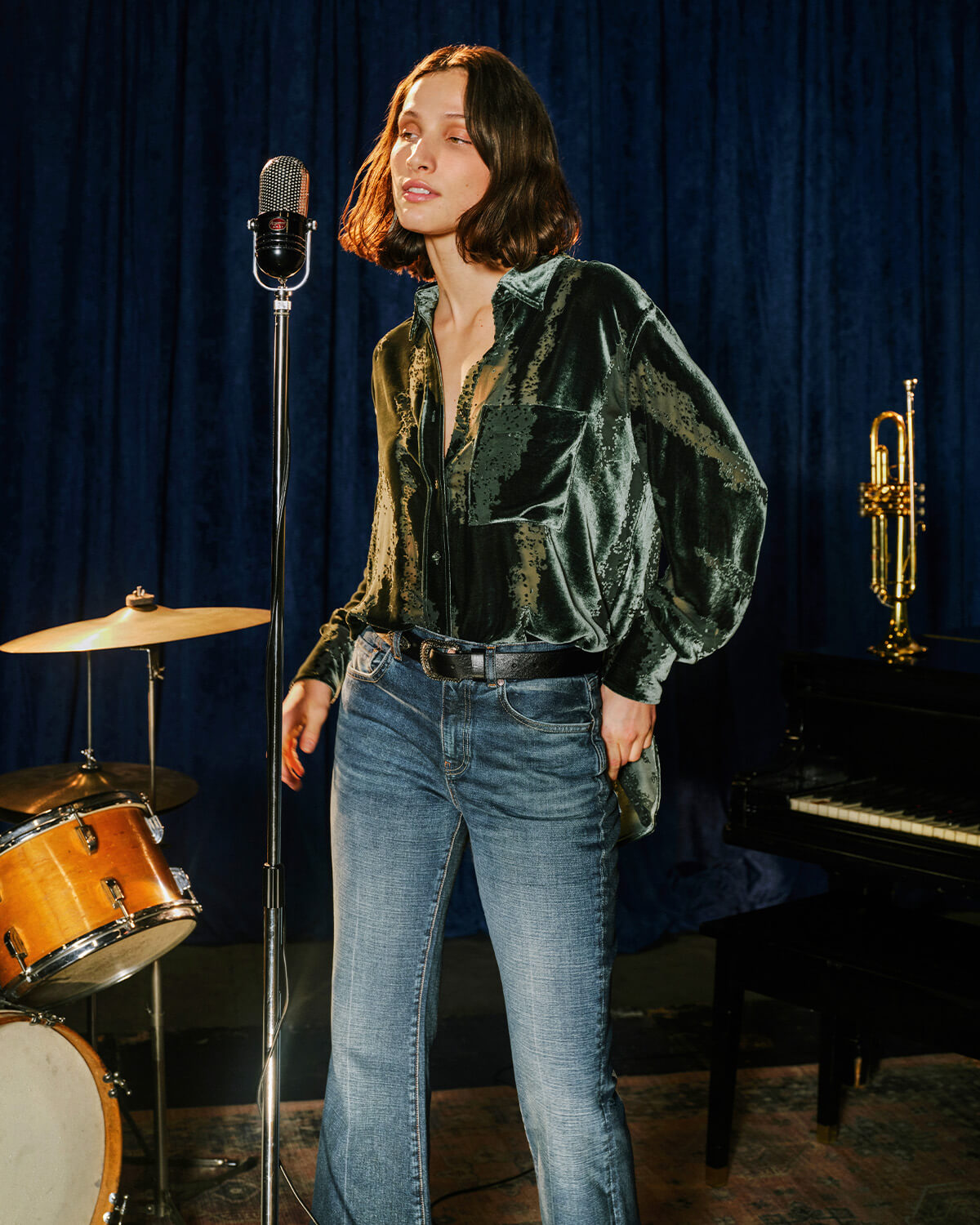 A woman stands in a studio setting wearing a dark green velvet long-sleeve blouse with a subtle tonal pattern and blue jeans. She is positioned in front of a vintage microphone, with a drum set, piano, and trumpet visible against a dark blue curtain backdrop.
