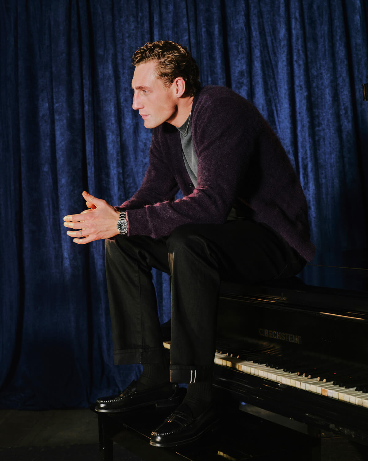 A man in a dark purple knit cardigan, black trousers, and black dress shoes sits on a black grand piano against a dark blue velvet curtain backdrop.