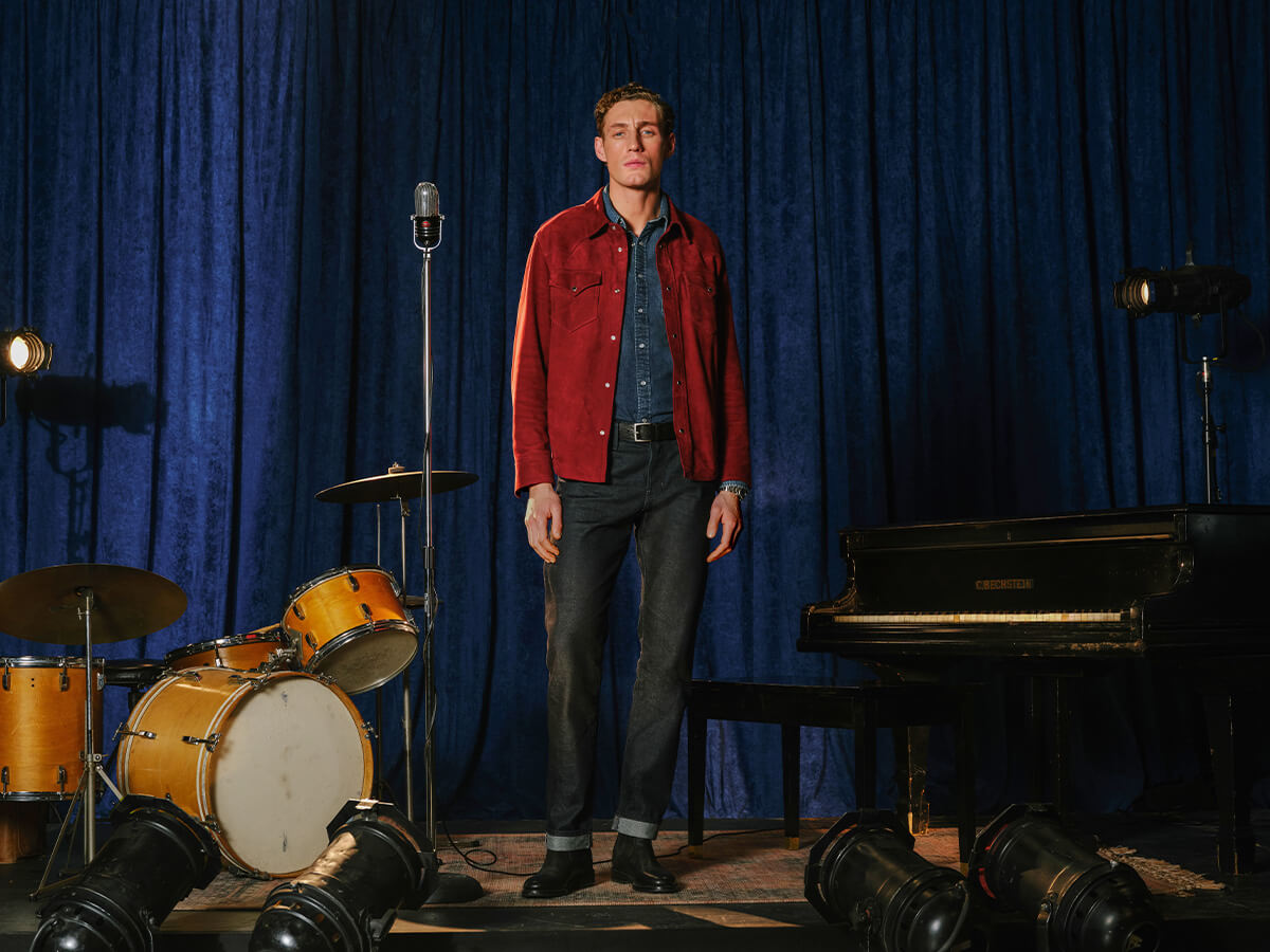 A man stands on a stage-like set with a blue velvet curtain backdrop, wearing a red button-up jacket over a blue shirt and dark jeans. A microphone, drum set, piano, and stage lights are visible around him.