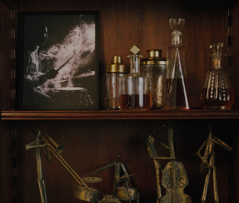 A framed black and white photograph of a drummer hanging on a dark wood shelf next to decorative glass bottles and decanters, with metallic percussion-themed sculptures below.