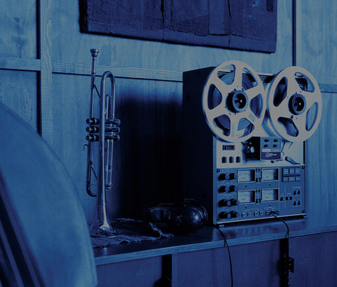 A vintage reel-to-reel tape recorder sits on a table next to a trumpet in a dimly lit room with a blue tint.