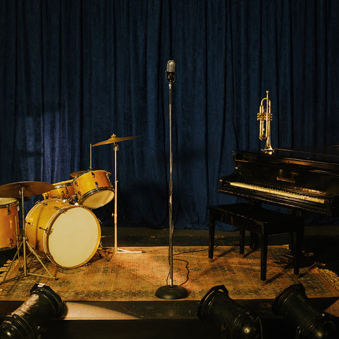 A dark stage setup with a blue curtain backdrop, featuring a drum set on the left, a vintage microphone on a stand in the center, and a black grand piano with a trumpet on the right.