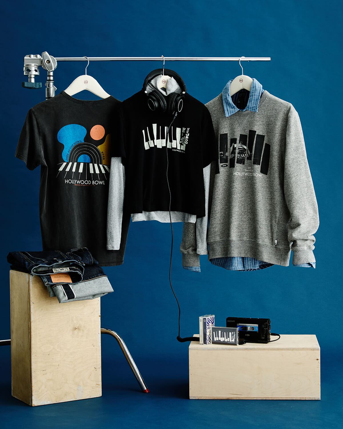 Three graphic hoodies and a gray sweatshirt from the brand homescapewear hang on a metal clothing rack against a dark blue studio backdrop, with folded jeans on a wooden block and small accessories on another block below.