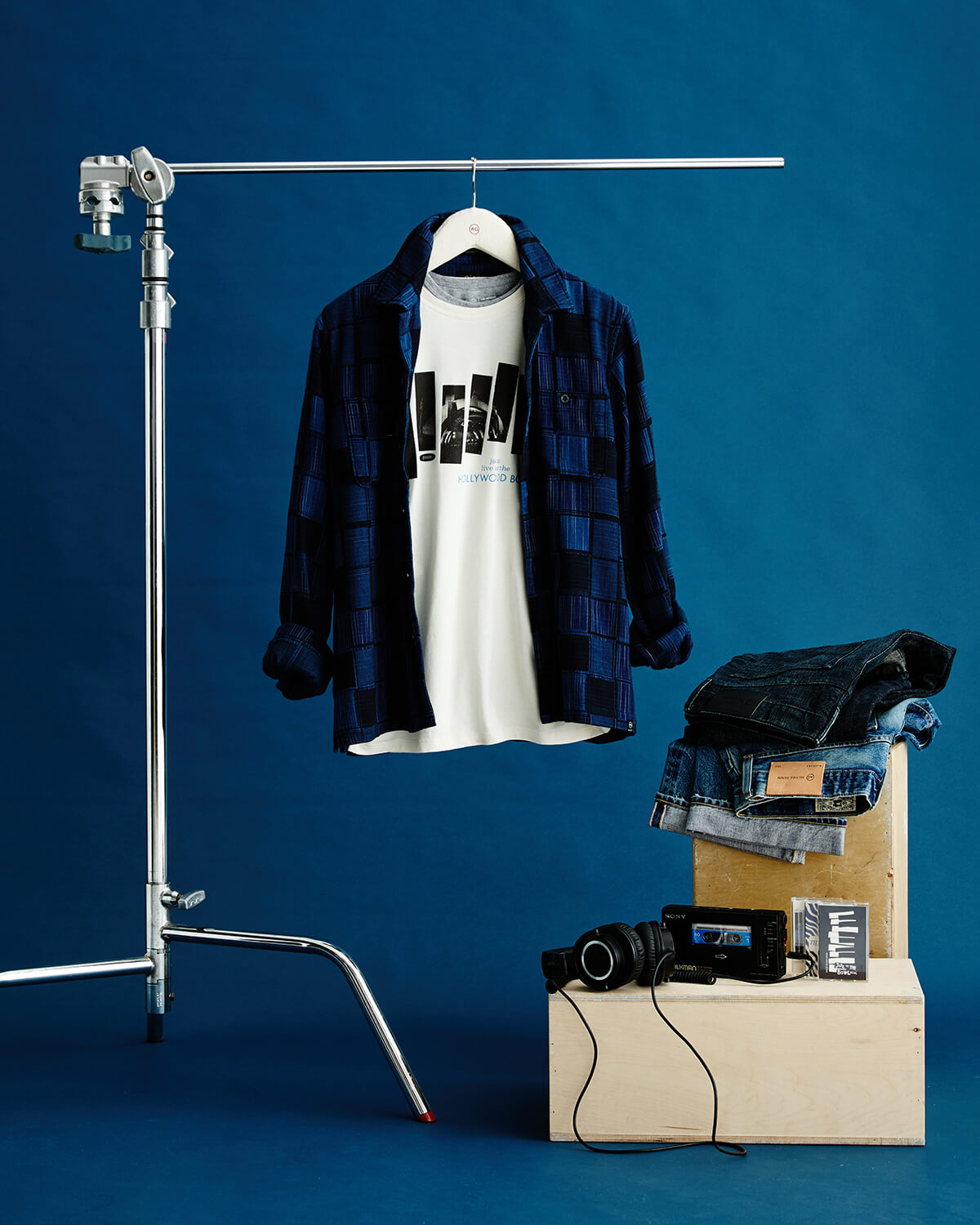 A styled fashion setup against a deep blue backdrop featuring a navy blue plaid overshirt layered over a white graphic T-shirt hanging on a hanger stand, with folded denim and a camera on a wooden block beside it.