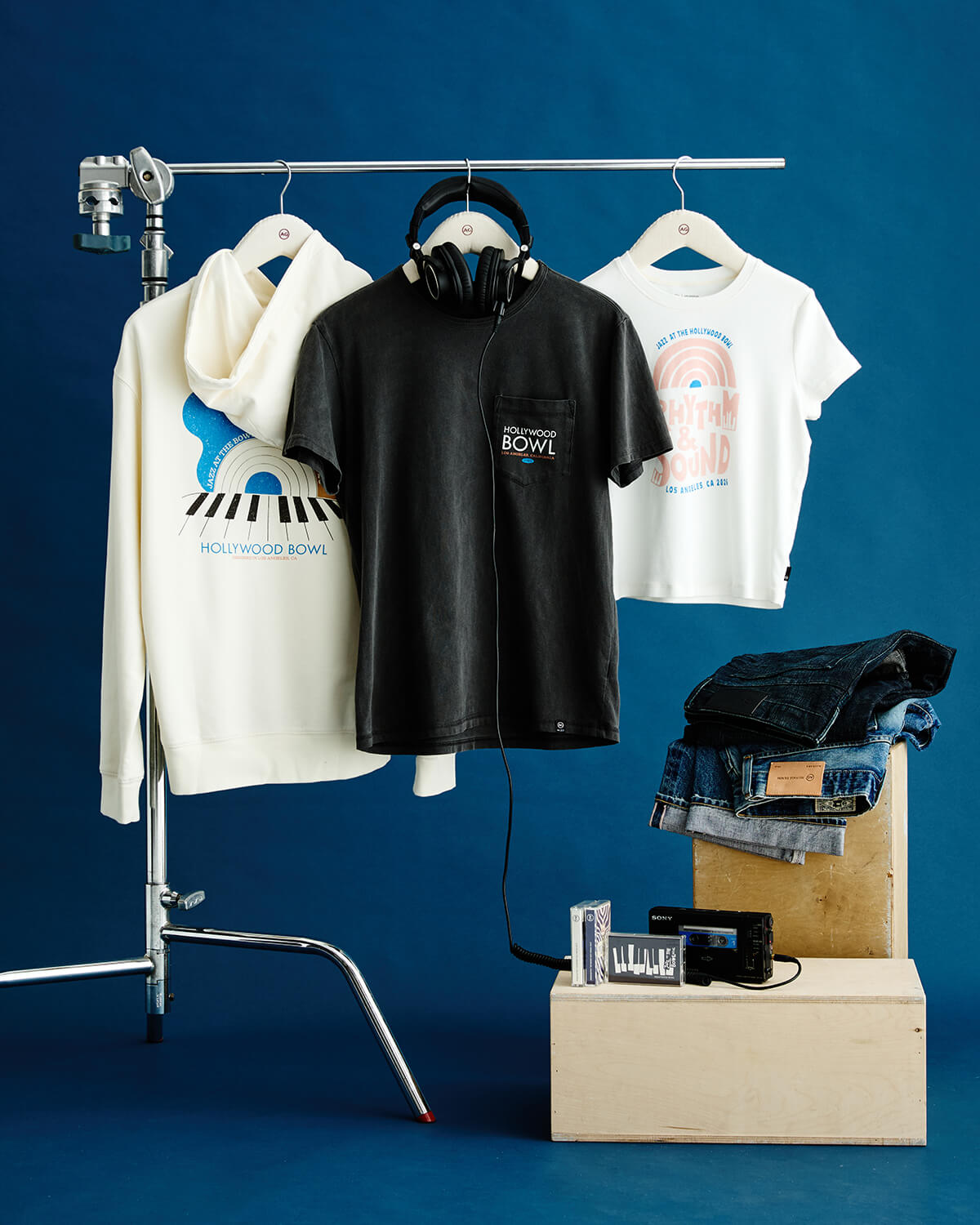 A styled apparel display on a blue studio background featuring a cream hoodie, a black zip-front t-shirt with a chest pocket and 'HOLIC SOUND' logo, and a white graphic t-shirt, all hanging on a stand. A pair of black headphones hangs from the shirt collar, with folded jeans and small accessories arranged on wooden blocks below.