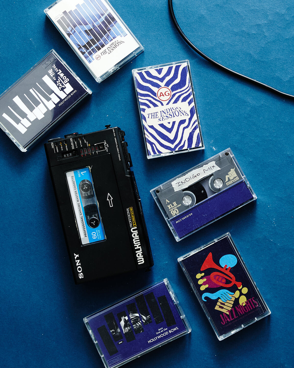 A styled flat-lay of vintage cassette tapes and a Sony Walkman tape deck on a textured blue background. The collection includes various cassette cases with colorful covers, including zebra print, piano keys, and jazz charts, alongside the black Sony Walkman cassette deck.